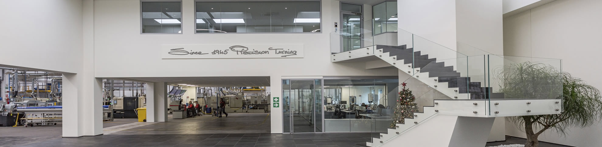 Interno dell'azienda Eredi Baitelli, che mostra la zona uffici moderna e ben progettata, con una vista sul laboratorio di torneria. L'immagine evidenzia un ambiente di lavoro pulito e sicuro, con un design contemporaneo che promuove il benessere dei dipendenti e l'efficienza produttiva. La zona è ben illuminata, con un albero ornamentale e un albero di Natale, creando un'atmosfera accogliente e innovativa.